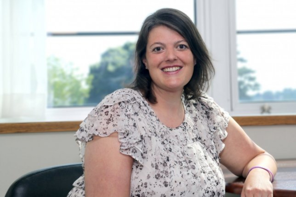 A smiling woman with shoulder-length dark hair sits indoors by a window, wearing a light floral blouse, with natural light illuminating her face.