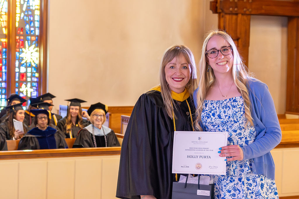 Two women stand together inside a church during a graduation ceremony. One wears academic regalia, while the other holds a certificate and a black gift bag. Graduates in caps and gowns are seated in the background, with stained-glass windows and wooden architectural details visible.