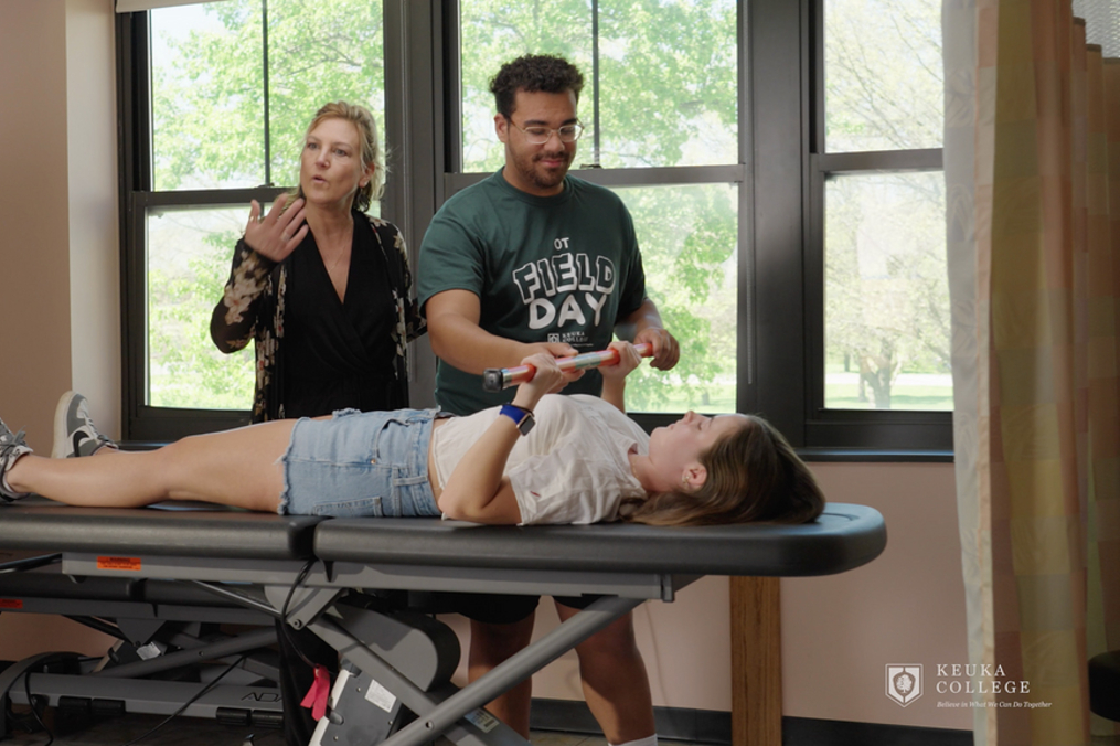 A student lies on a medical examination table while holding a piece of equipment during a hands-on healthcare training activity. A student wearing a “Field Day” shirt assists, while an instructor stands nearby and explains the procedure. Large windows provide a view of trees outside.