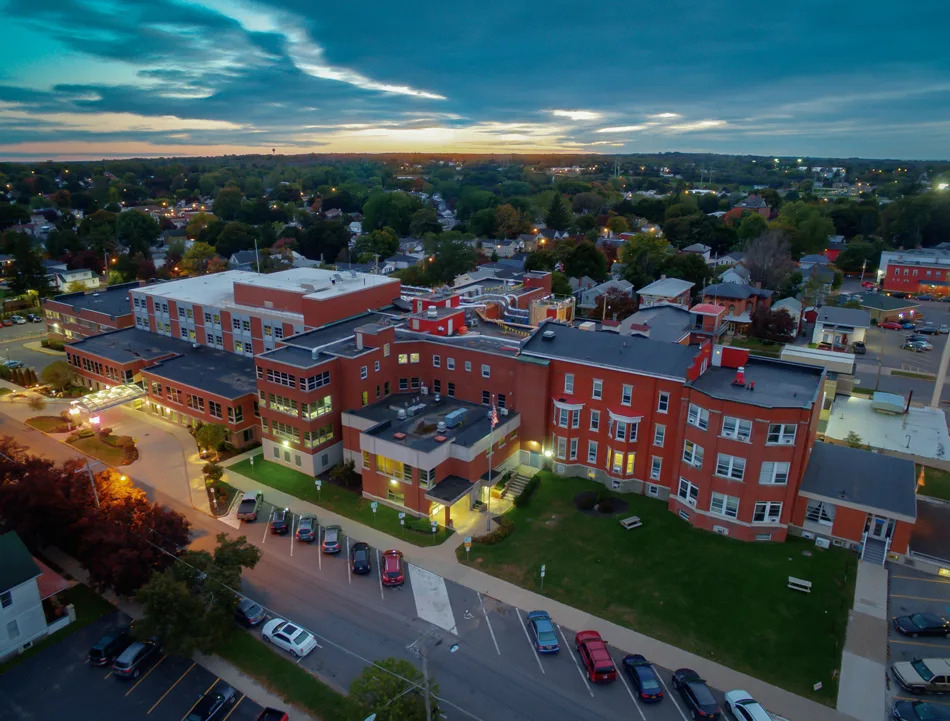 Aerial view of a large red-brick institutional building complex at sunset, surrounded by a small town with tree-lined streets, parked cars, and low-rise houses.