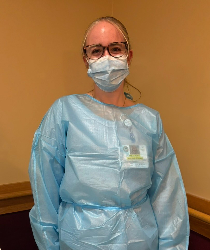 A healthcare worker wearing glasses and a surgical mask stands facing the camera in a clinical setting. They are dressed in a light blue disposable protective gown with an identification badge attached to the front.