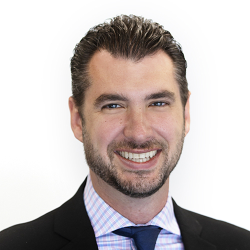 Headshot of a man with short dark hair and a trimmed beard, smiling at the camera. He is wearing a dark suit jacket, a light checkered dress shirt, and a blue tie against a plain light background.